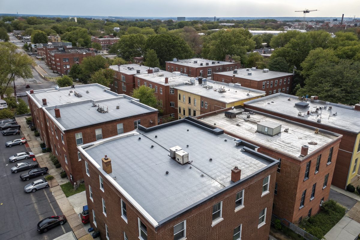 Aerial view of brick apartment blocks with gray flat roofs, rooftop units and a tree-lined street below