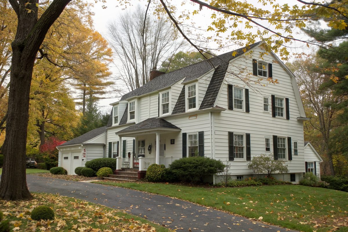 A white gambrel-roof colonial with dark shingles, shutters and a driveway under autumn trees